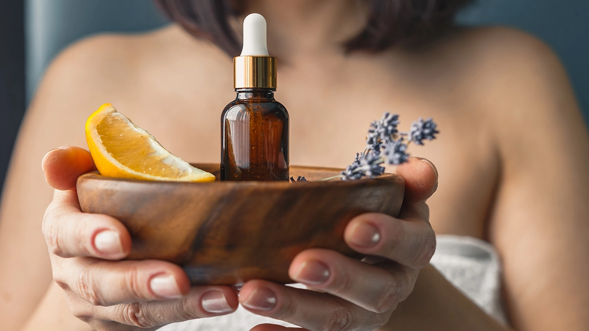 A woman holds a wooden bowl with an essential oil bottle, orange, and lavender, symbolizing aromatherapy benefits for energy, relaxation, and clarity.