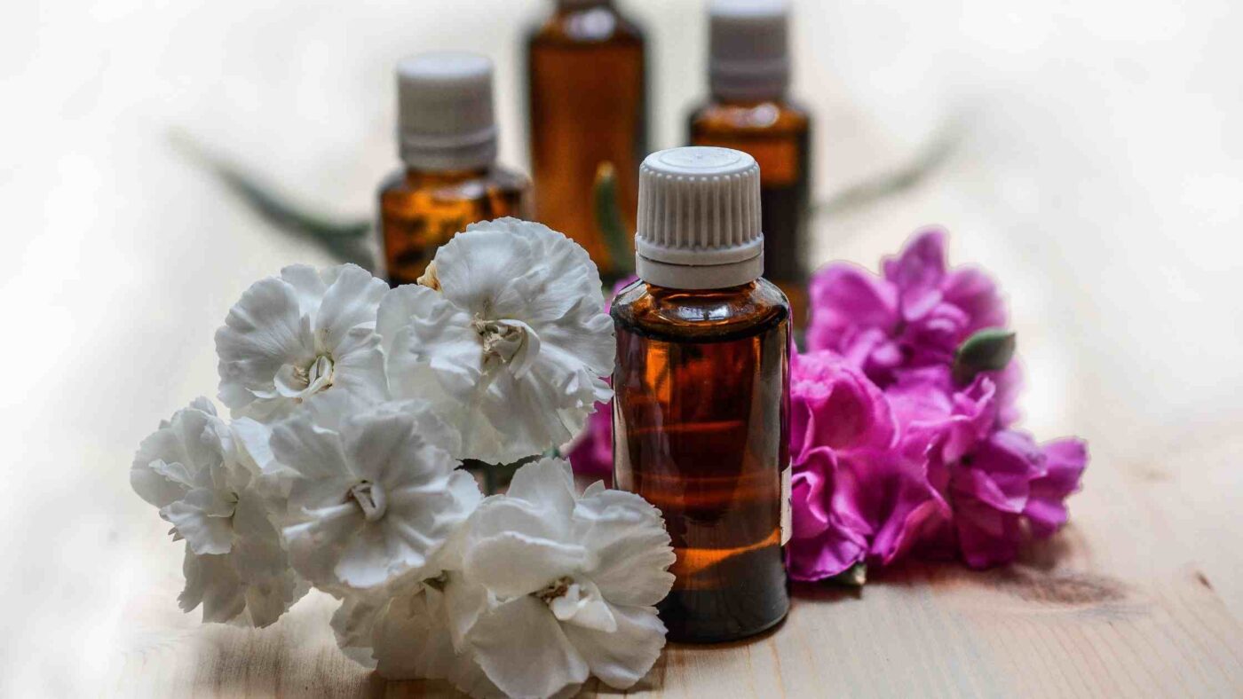 The image shows amber bottles of essential oils surrounded by white and pink flowers on a wooden surface, representing the concept of diffusing colloidal silver and essential oils together.