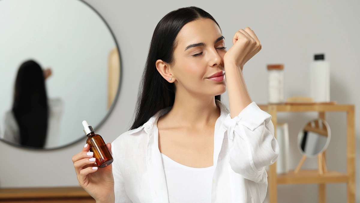 A person holding a bottle of essential oil with a dropper, standing in front of a mirror in a room with neutral colors. The individual’s reflection is visible in the mirror, creating a sense of self-awareness and connection.