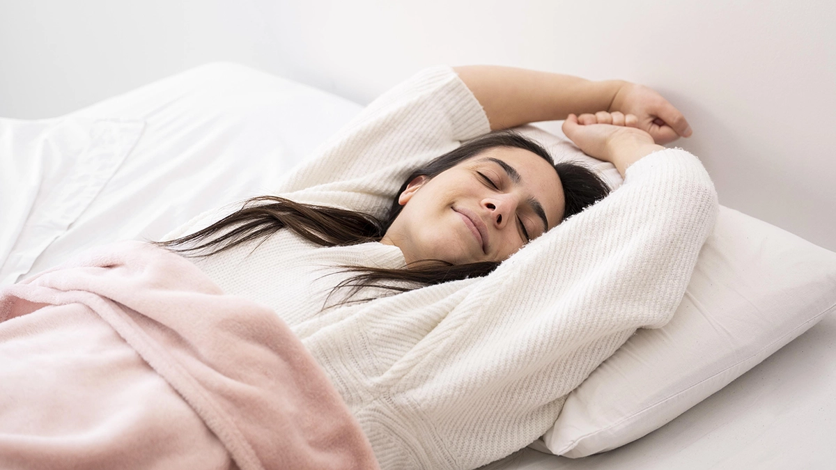 A woman stretches contentedly in bed, wrapped in a soft pink blanket, looking relaxed and peaceful