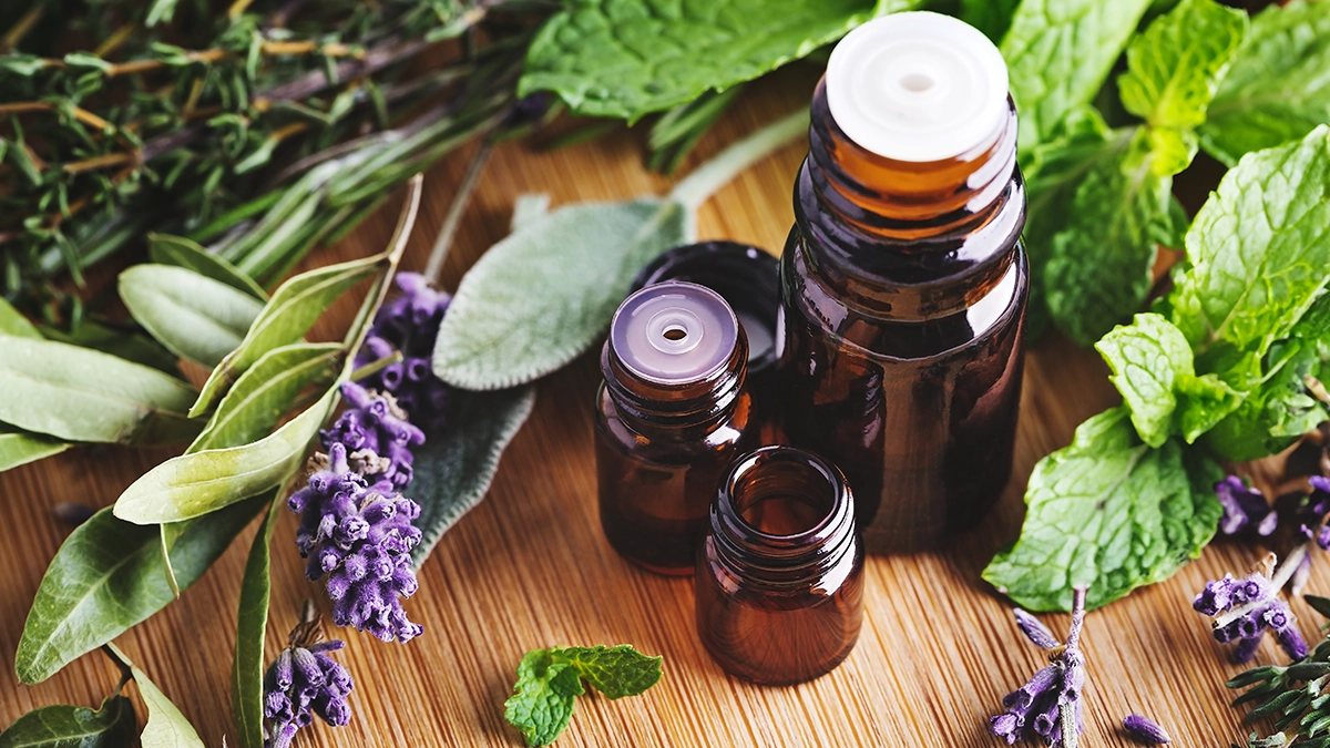 Brown glass bottles of essential oils surrounded by fresh herbs and flowers, including lavender, peppermint, and eucalyptus