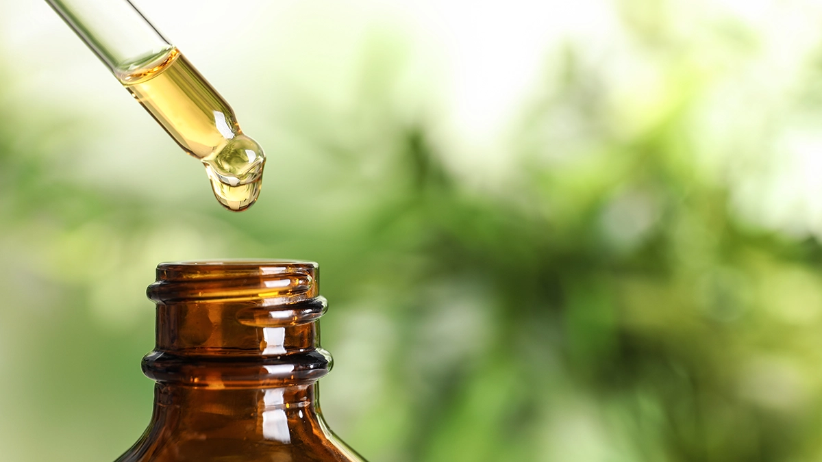 A dropper dispensing essential oil into an amber glass bottle, with a blurred natural background