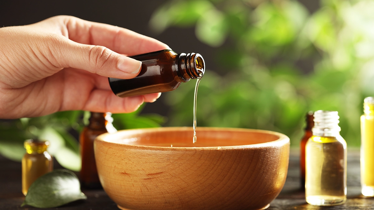 A hand carefully pours essential oil from a brown bottle into a wooden bowl, surrounded by other essential oil bottles