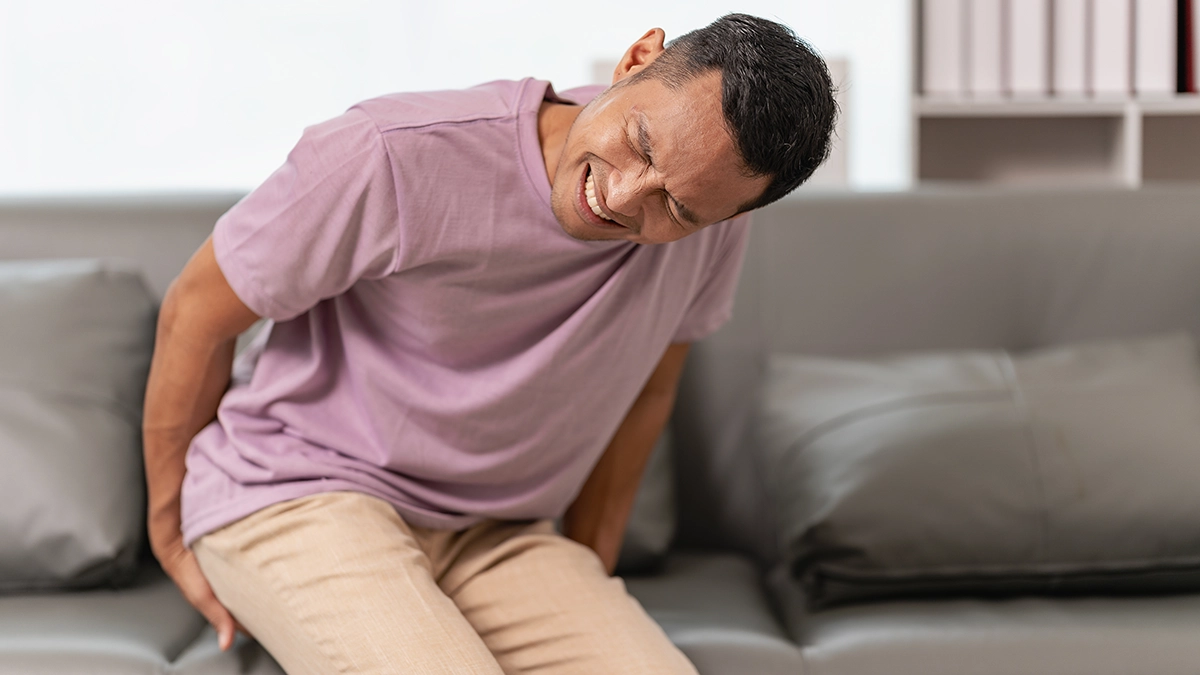 A man in a purple shirt sits on a sofa, wincing in pain while holding his lower back, indicating discomfort from hemorrhoids