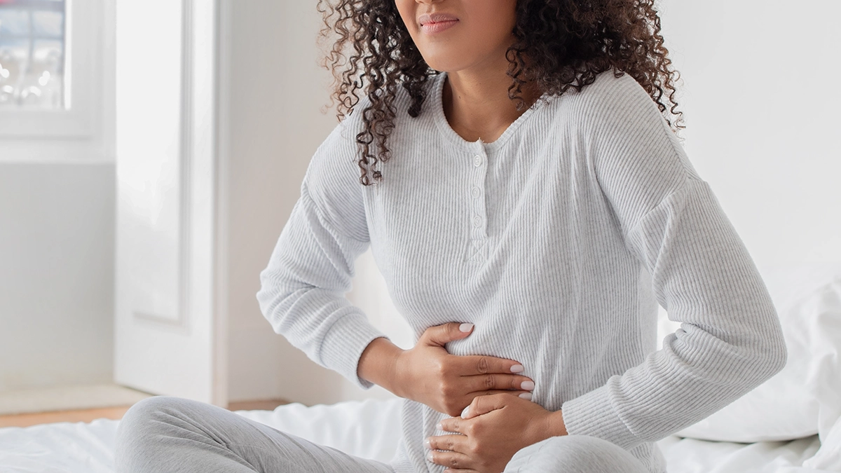 A woman sitting on a bed holding her stomach, appearing in discomfort, possibly due to digestive issues or diarrhea