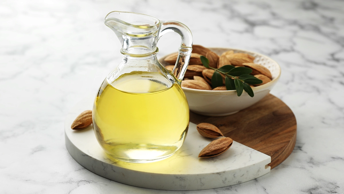 A glass jug of carrier oil beside almonds highlights its role in moisturizing and nourishing the skin