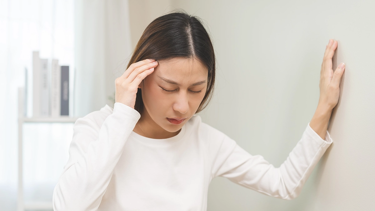 A woman experiencing vertigo, holding her head with one hand and steadying herself against a wall, eyes closed in discomfort