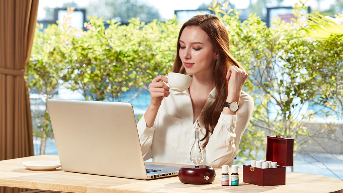 A woman relaxes with tea, a laptop, and an Organic Aromas diffuser with essential oils for calming support