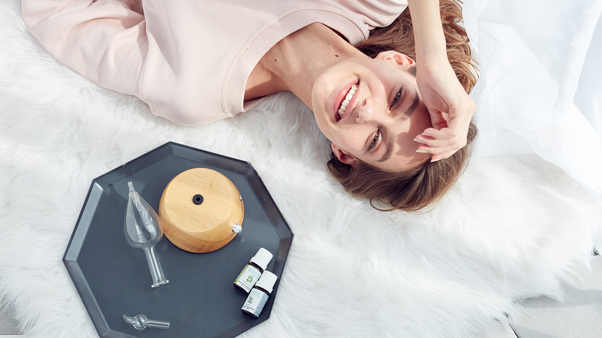 A woman relaxing on a white rug with a wooden diffuser, glass attachments, and essential oils on a tray nearby