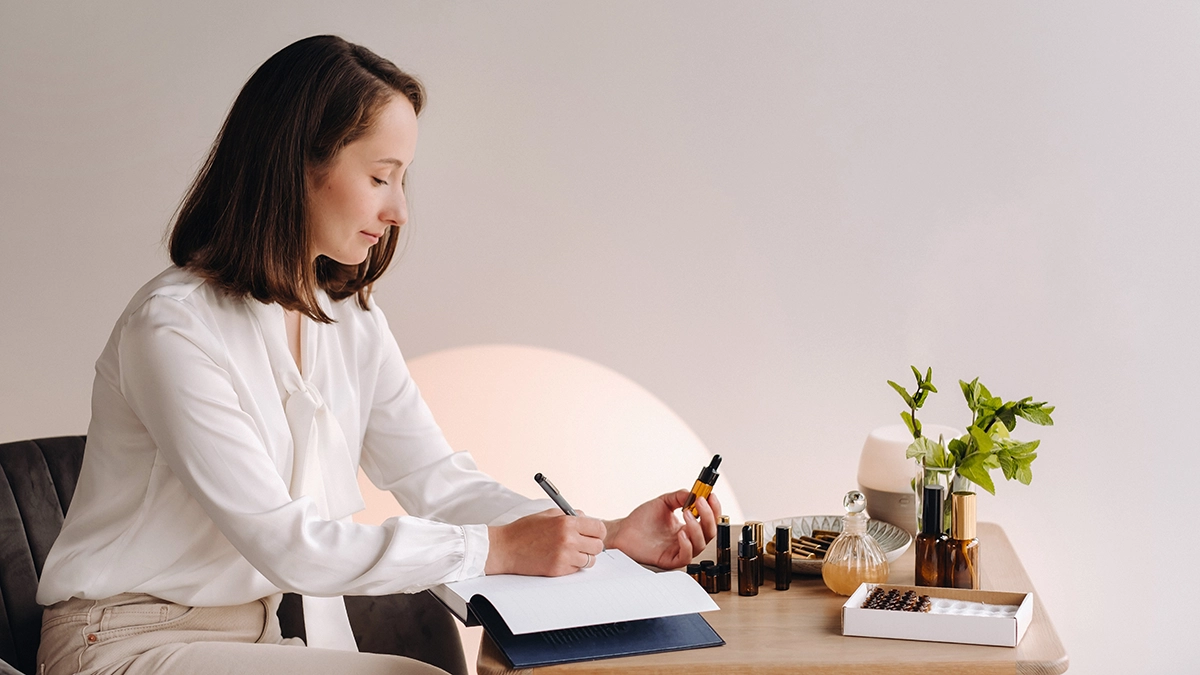 A woman writes while holding an essential oil dropper, surrounded by bottles and plants. Oils like rosemary, peppermint, and lemon enhance focus and memory.