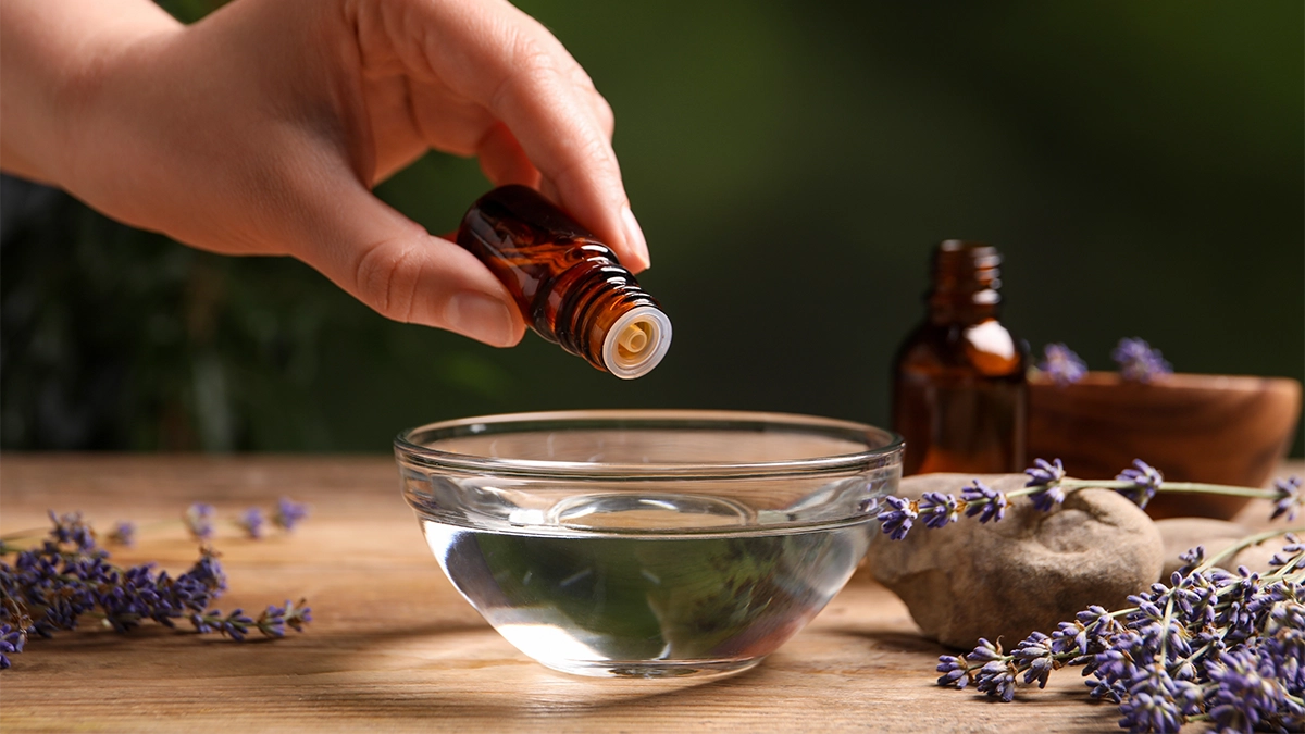 Hand adding lavender essential oil drops into a bowl of water, illustrating DIY topical and aromatic sleep aids with lavender