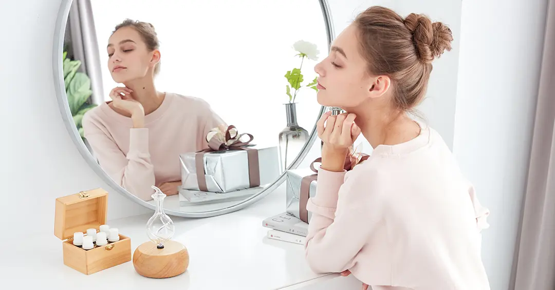Woman applying essential oil to her neck, sitting at a vanity with a mirror, wooden diffuser, and essential oil bottles.