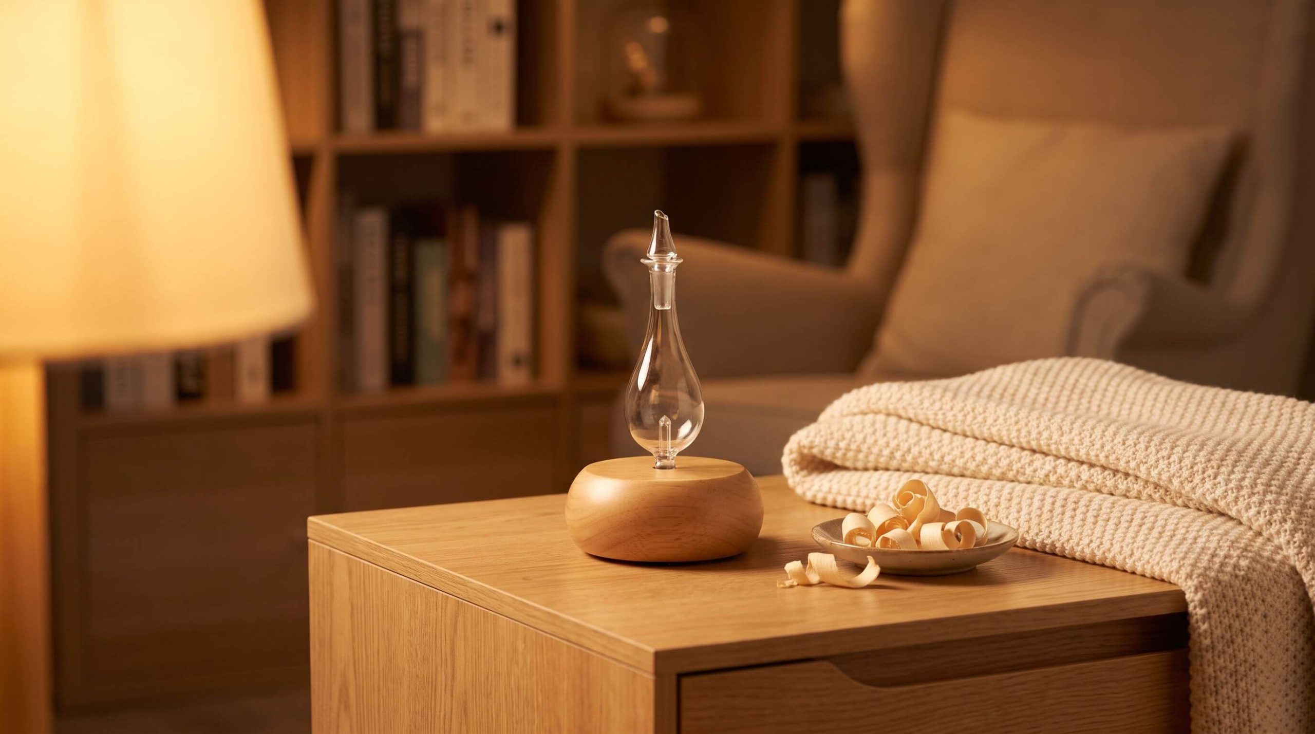 Handcrafted wood and glass Nebulizing DiffuserĀ® at rest on an oak side table in a warm reading nook beside pale sandalwood shavings